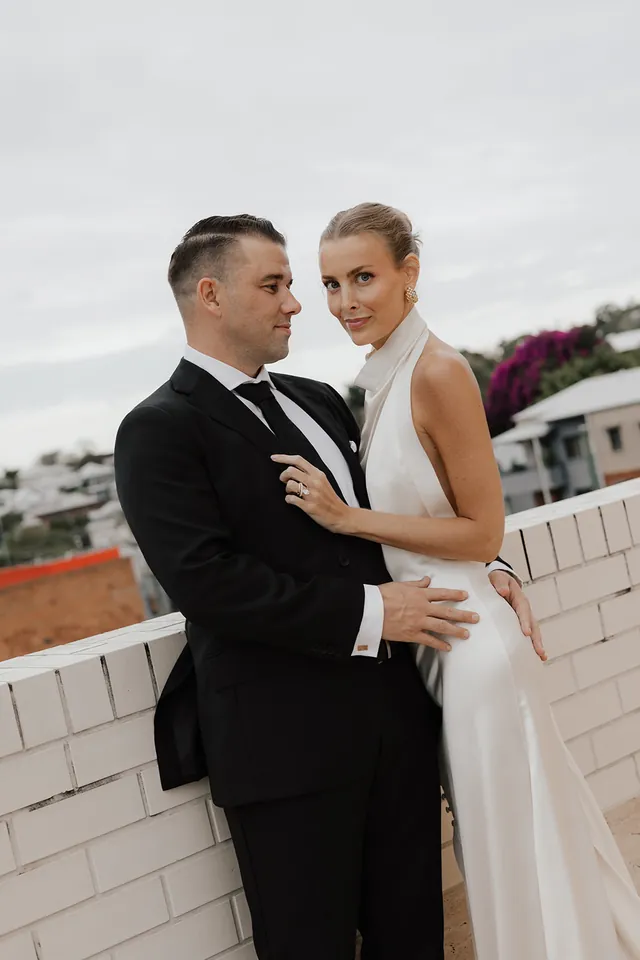 Clare and Matt embracing beside a white brick terrace wall and the Brisbane skyline.