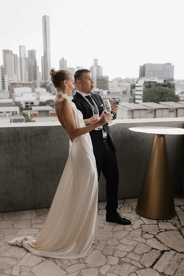 Clare and Matt holding champagne on The Calile terrace with Brisbane behind them.